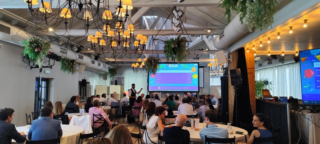 Gamificación para eventos: salón con los equipos sentados por mesas y el marcador del Concurso de TV en la pantalla