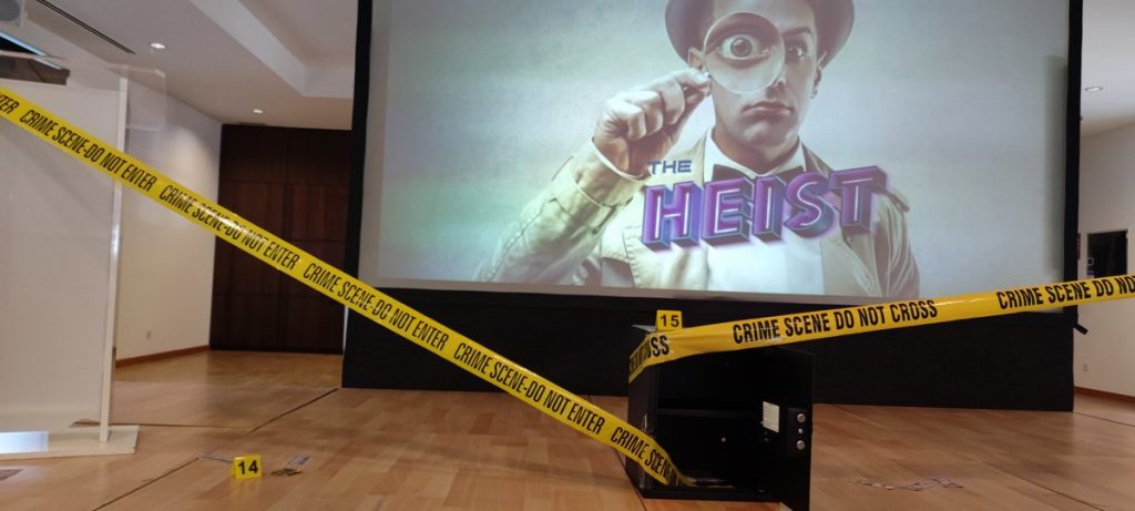 Dinámicas de trabajo en equipo presenciales: pantalla con la portada de The Heist y cinta de escena del crimen en una sala de eventos